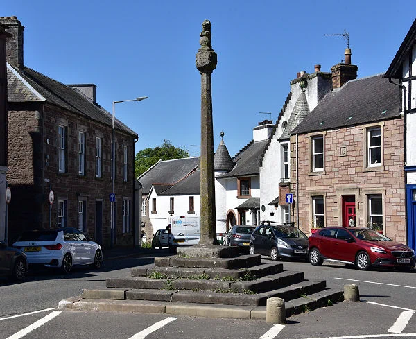 Today's 'Where in Scotland' image. It is a squarish image showing a mercat cross standing on a six-stepped base in the centre of the frame with roads both sides of it and, with white hatching, across the bottom of the frame. There are two-storey buildings both sides of the frame in stone, with a white building downhill from the cross in the background. The sky is blue.