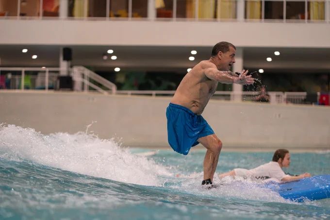 Man in blue swim trunks balances on an indoor wave machine while another person lies on a blue board behind him.
