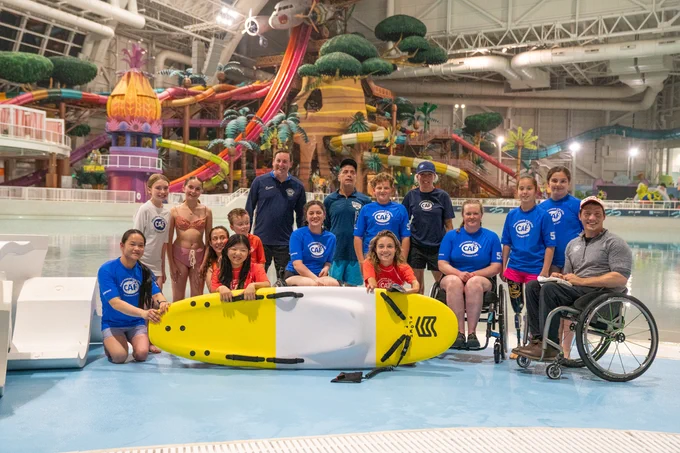 Group of adaptive athletes and coaches pose with a surfboard and wheelchairs at an indoor water park wave pool.