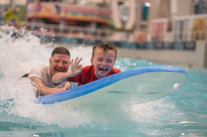 Smiling boy in a red shirt rides a surfboard indoors, making an OK sign, while an adult behind him laughs in the waves.
