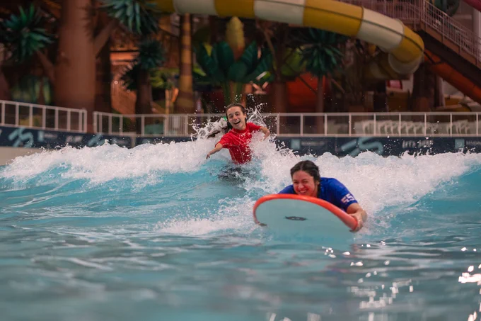 Two people ride an indoor wave pool, one on a bodyboard and one splashing behind, with water slides in the background.