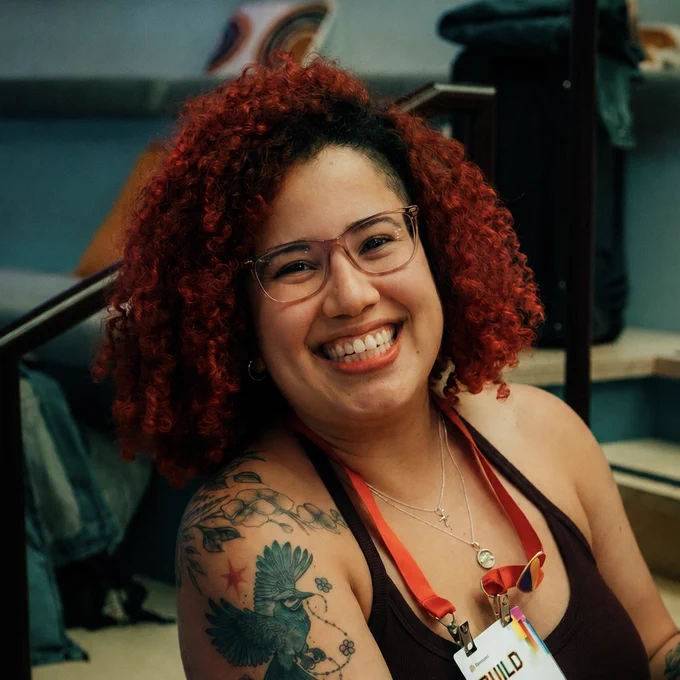A smiling individual with curly red hair and glasses displays a tattoo on her arm, wearing a tank top and a name badge.