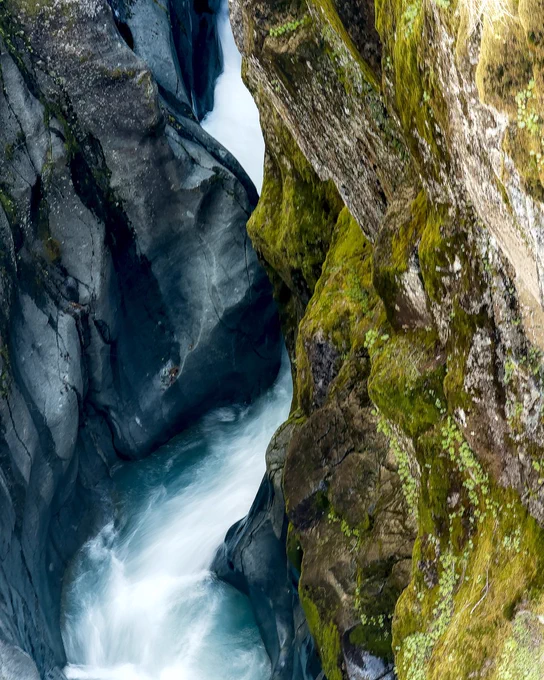 A vertical landscape photo looking down into a narrow rocky canyon where white water rushes between steep stone walls. The left side shows smooth, dark blue-gray rock shaped by flowing water, while the right side is covered with green moss, lichen, and textured brown stone. The water curves through the tight channel, creating soft white motion against the darker rock. The scene feels cool, powerful, and carved by time. The photo makes me feel drawn into the force and patience of moving water.