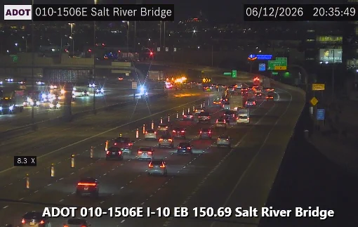 Traffic on a well-lit multi-lane highway, with cars and an illuminated overhead sign displaying "I-10" near Salt River Bridge. Orange traffic cones are set up along the left side. Overhead, the image displays the date, time, and location details, including ADOT (Arizona Department of Transportation).