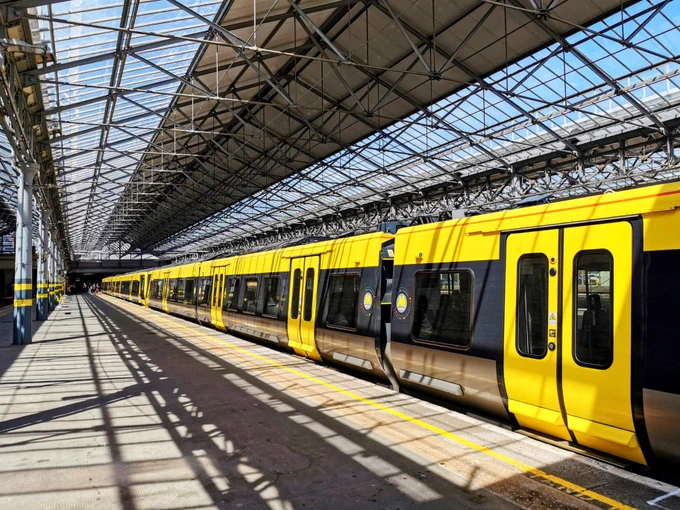 A class 777 Merseyrail train in the platform at Southport station under a large glass and steel roof with strong shadows cast by the supports.

The scene composition is of a train platform beneath a large, glass-covered roof with a yellow and black train parked along it. The train is in the centre of the image, and the platform stretches out from the foreground to the midground. The overhead structure, a large glass-covered roof, casts parallel shadows across the platform that is wide, paved and marked with yellow lines.
