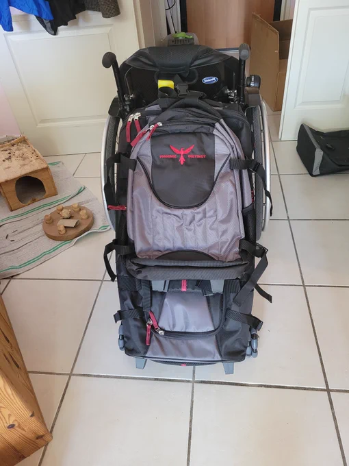 Un fauteuil roulant de dos auquel est accroché un grand sac de voyage avec des roues omnidirectionelles. Un sac à dos assorti est accroché sur le sac de voyage. 