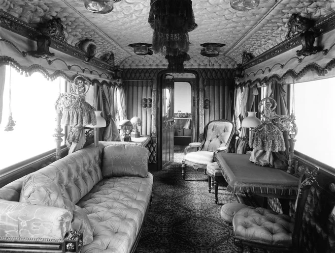 Victorian-era train carriage interior with ornate furnishings, upholstered sofas, intricate woodwork, and decorative lamps. The ceiling features elaborate patterns, and large windows are adorned with draped curtains.