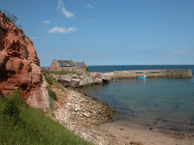 Cove near Cockburnspath. The image shows a coastal scene. There is an outcrop of exposed red rock on the left with a grassy bank this side of it and a stony beach to its right which leads into a blue sea. Beyond the rock are two cottages standing on the start of a stone jetty. There are three small boats moored in the harbour. The scene is in sunlight.