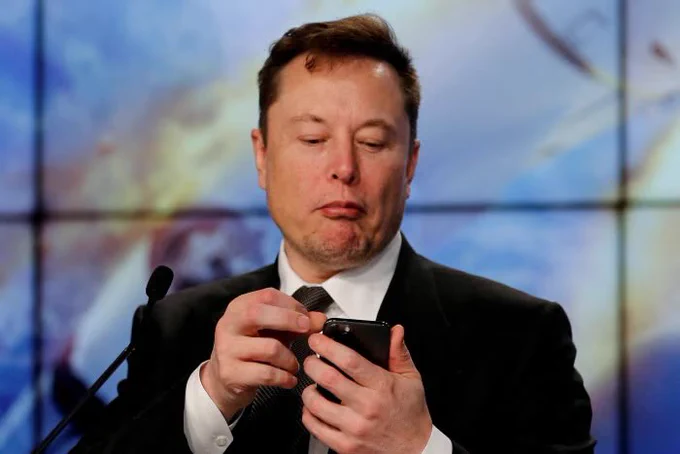 Elon Musk looking at his iPhone while wearing a suit for a press conference.
