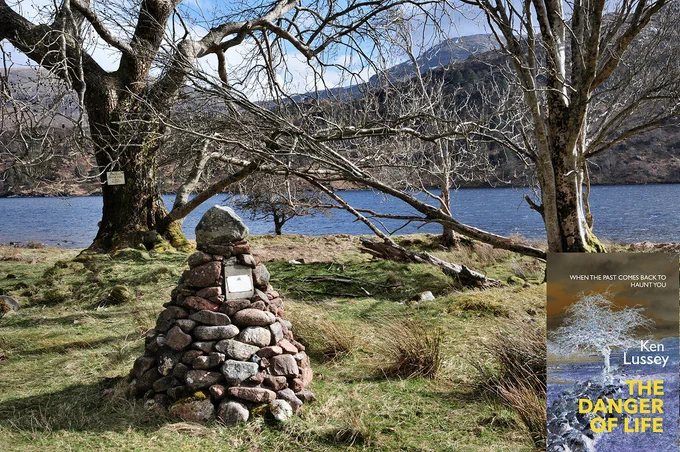 The image shows a stone cairn with a plaque in the lower left of the frame standing on rough grassy land with some bare trees behind it. One of the trees also has a plaque on it. Beyond the trees is the water of a loch that stretches across the centre of the frame. Beyond the loch the land rises to hills. The scene is in sunlight. The front cover of ‘The Danger of Life’ is shown in the bottom right corner.