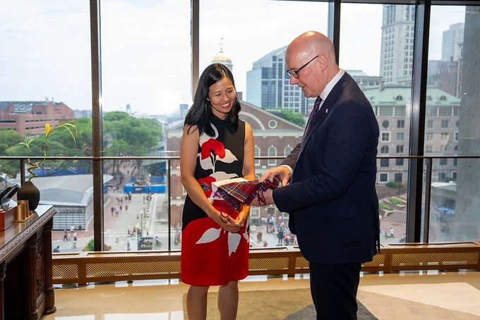 The Mayor of Boston in a red and black dress looks at a tartan scarf held by the First Minister, who is wearing a suit. The two stand inside a room with large windows overlooking a cityscape.