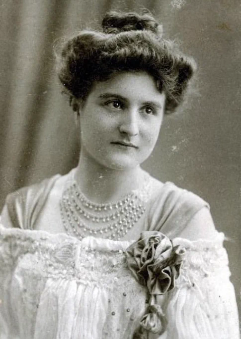 Vintage black-and-white portrait of a woman dressed in an elegant, early 20th-century outfit with detailed embellishments and multiple strands of pearls.