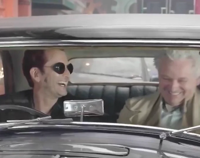 david tennant and michael sheen in the bentley laughing, looking happy. they are dressed as crowley and aziraphale. it’s them