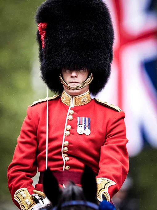 A ceremonial guard in a red uniform and tall black bearskin hat rides on horseback during a military parade, with a British flag in the background. Photo by UK MOD © Crown copyright 2026.