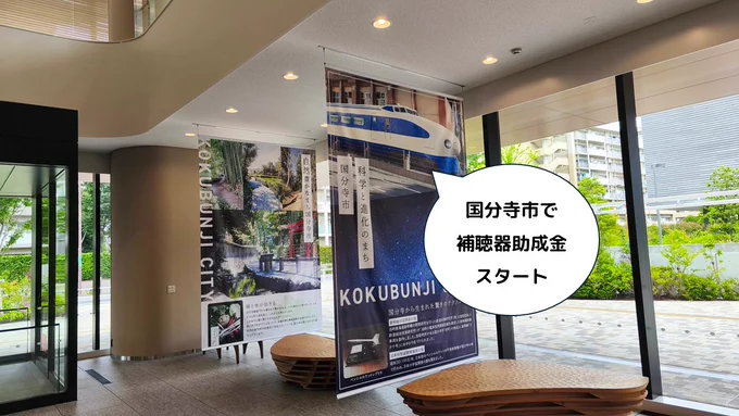 国分寺市役所のエントランスホールの写真。正面に「科学と進化のまち 国分寺市 KOKUBUNJI」と書かれた0系新幹線の大型垂れ幕が下がり、左手には「自然豊かなまち 国分寺市 KOKUBUNJI CITY」の緑の写真パネルが並んでいます。右側はガラス張りで、外の街並みと緑が見えています。画像右側に吹き出しがあり「国分寺市で補聴器助成金スタート」と書かれています。国分寺市では令和8年7月1日から、満65歳以上の方を対象に高齢者の補聴器購入費の助成（上限4万円）が始まります。申請は購入前に市役所2階の地域包括ケア課で行います。