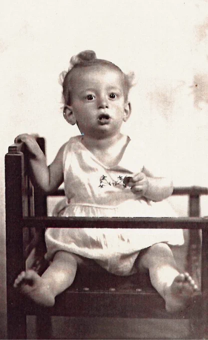 A black and white photo of a baby sitting upright in a wooden chair, holding a small object and looking surprised.