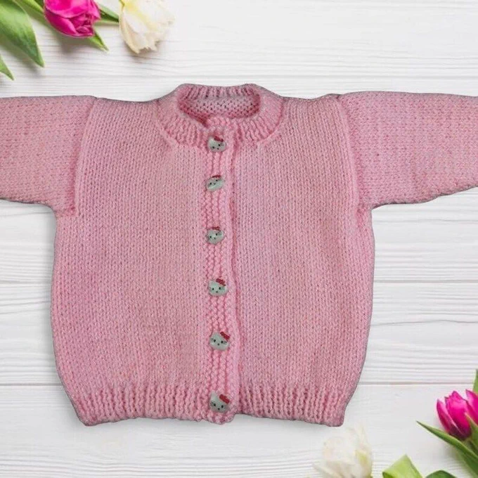 Soft pink knitted cardigan laid flat on whitewashed wooden planks. Features a simple stockinette stitch texture, a round neckline, and long sleeves. Fastened with a row of small decorative buttons down the front. Ribbed detailing at the hem and cuffs. Pink and white tulips with green leaves frame the top-left and bottom-right corners. Bright lighting highlights the smooth, even knit and gentle colour.
