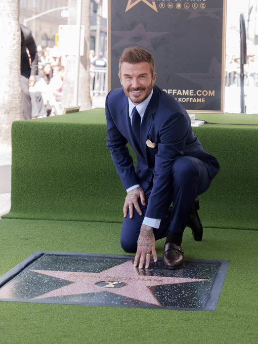 Sir David Beckham in a navy suit kneeling and touching a star on the Hollywood Walk of Fame during a ceremony.