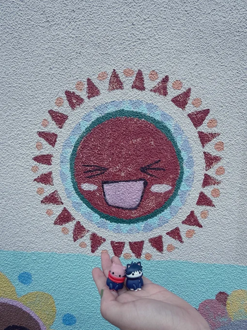 壁に描かれた笑顔の太陽を背景に撮った、猫としてデフォルメされた、いたどり、ふしぐろの小さいフィギュア