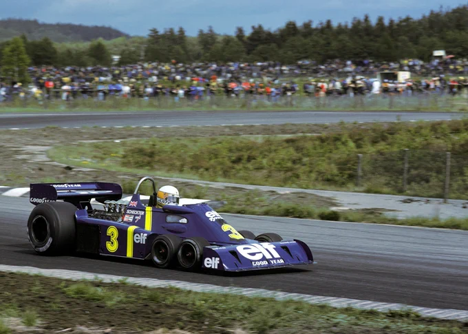 In the bottom left of frame, Jody Scheckter is on track facing rightwards in his blue and yellow Elf sponsored Tyrrell P34, which has two large rear wheels and four small front wheels. The track curves back out of shot and back in around a long corner, with spectators gathered on the outside of the track in the background. 1976 Swedish Grand Prix, David Phipps/Sutton Images