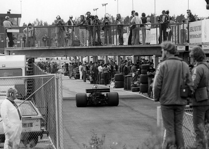 A black and white photo of Jody Scheckter's Tyrrell P34 being driven away from the camera as it drives down pit lane. A small, low footbridge passes over pit lane at the height of the single storey garages; a number of photographers stand taking photos of the pit area. 1976 Swedish Grand Prix, David Phipps/Sutton Images