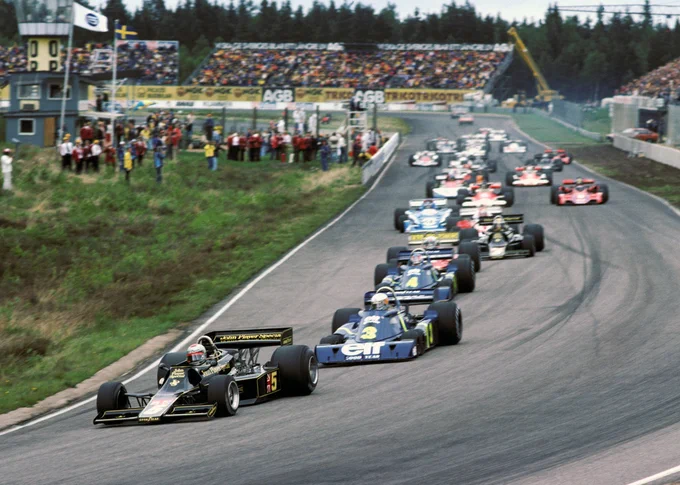 A view from turn one of the start, looking back down the track; in the bottom left of frame turning right, Mario Andretti leads in his black and gold JPS Lotus 77, with the two blue and yellow Elf sponsored Tyrrell P34 cars of Jody Scheckter and Patrick Depailler close behind. The rest of the pack follows behind. 1976 Swedish Grand Prix, David Phipps/Sutton Images