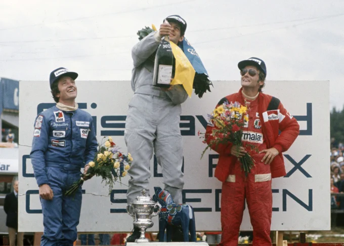 The podium; Jody Scheckter stands in the middle on the highest step in pale blue race overalls, laurels around his neck as he pops his champagne cork straight up into the air. On the left of frame, Patrick Depailler watches and smiles, wearing darker blue race overalls and holding a small bunch of flowers. On the right of frame Niki Lauda also watches while smiling, a hand on his hip while holding a bunch of flowers. He wears a red race suit, a red jacket with black striped shoulders and aviator sunglasses. All wear dark blue Goodyear caps. 1976 Swedish Grand Prix, LAT Images