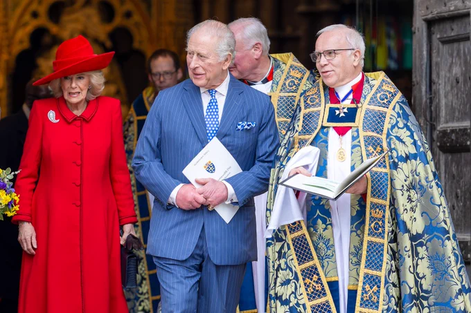 HM The King, wearing a blue pinstriped suit, leaving Westminster Abbey following a service. He is accompanied by HM The Queen, wearing a vibrant red coat and matching hat, with the Dean of Westminster standing to their left wearing a blue a gold cope.