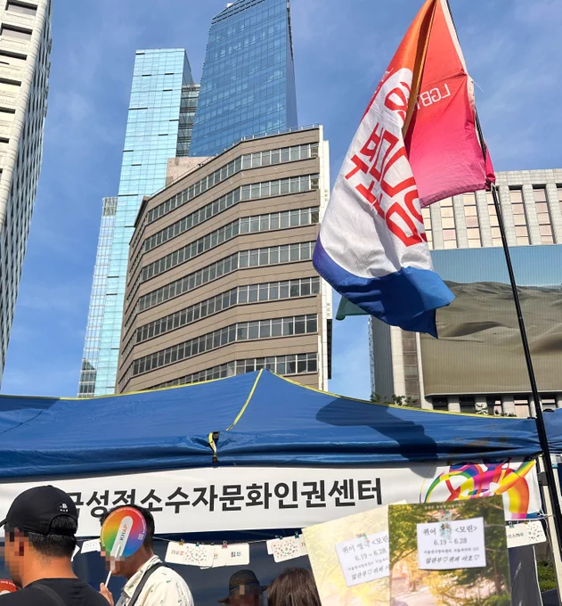 2026 서울퀴어퍼레이드 부스 중, 한국성적소수자문화인권센터 부스 앞에서 포스터를 들고 촬영한 사진이다. 오른쪽에는 성소수자부모모임 깃발이 바람에 나부끼고 있다.