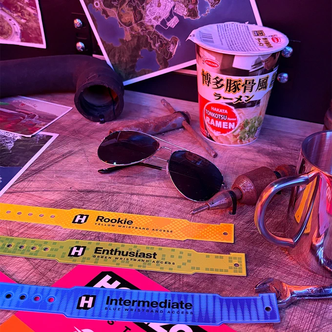 A close-up of the workbench tabletop showing sunglasses, ramen, and Forza Horizon 6 Rookie, Enthusiast, and Intermediate wristbands.
