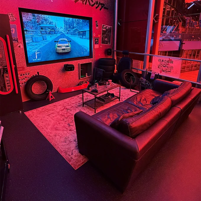 Another angle of the workshop installation at the event showing a lounging area with a black leather couch, rug, and tyres in the corner. Forza Horizon 6 plays on the flat-screen TV.