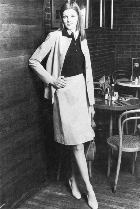 A woman is standing in a cafe setting, leaning against a wooden wall. She is wearing a tailored jacket, a skirt, and holding a handbag. In the background, two small American flags and some tableware are visible on a table with wooden chairs.
