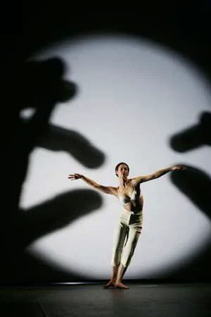 A contemporary dancer performs under a bright spotlight while large, blurry hand silhouettes frame her from both sides, creating a surreal forced-perspective effect.