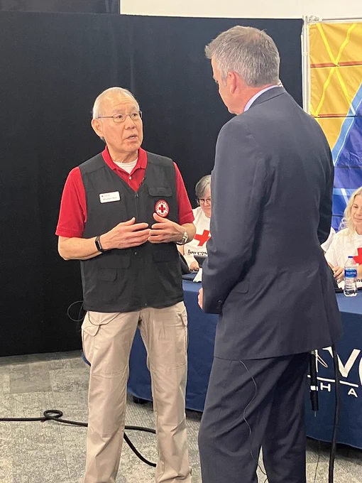 An individual with a red shirt and Red Cross vest speaks with a person in a suit. In the background, there are people seated at a table with Red Cross materials.