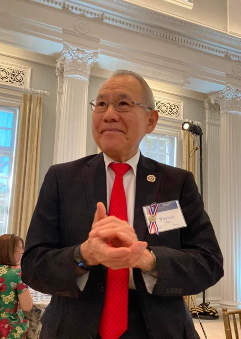 A person is standing in a room with ornate white columns and large windows. They are wearing a dark suit, red tie, and a name badge. The individual is smiling and appears to be at an event or function.