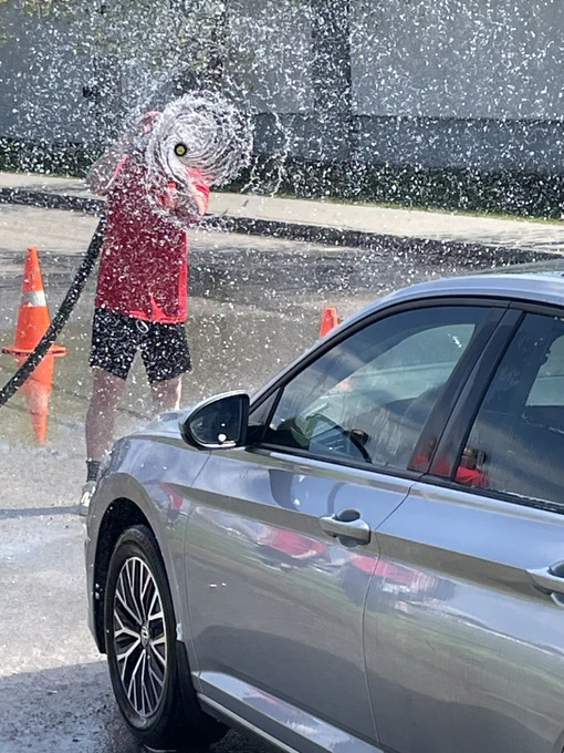 A Fire Cadet in a red shirt sprays a water hose at the front of a silver sedan.