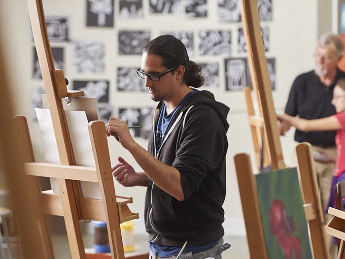 A focused artist works on a painting in a studio filled with easels and paintings. The atmosphere is creative and studious.