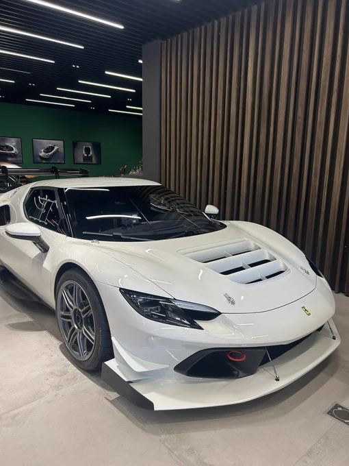 A white Ferrari 296 Challenge track car displayed on a low plinth inside a modern Ferrari dealership or atelier. The car features an aggressive front splitter, a louvered engine cover with cooling vents, gloss silver multi-spoke forged wheels on Pirelli slicks, a roll cage visible through the windshield, and the yellow Ferrari shield on the front fender. Behind it, a green accent wall displays framed Ferrari prints, a slatted wood feature wall sits to the right, and linear ceiling lighting runs overhead. A second race car with a rear wing is partially visible to the left.