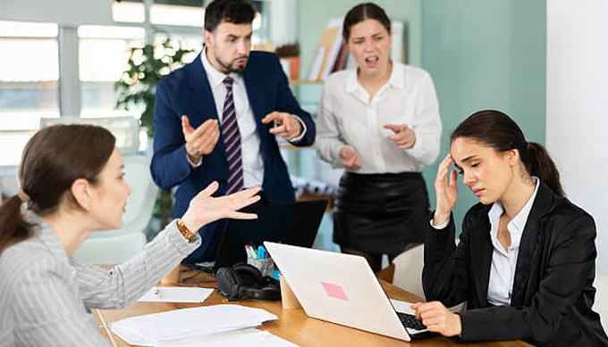 Office workplace conflict scene showing stressed employees in a heated discussion around a desk, with one worker frustrated at a laptop while others argue and gesture, illustrating workplace tension, miscommunication, and conflict management challenges in a corporate environment.