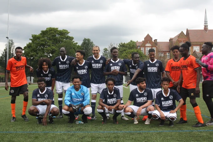 A diverse football team in blue and orange kits posing on a pitch, with a brick building in the background.