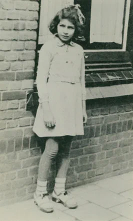 A historical black-and-white photo of a young girl standing alone in front of a brick building, dressed in a buttoned dress with a collar, looking into the camera.

