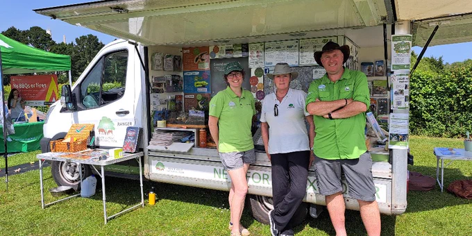 A mobile information van for the New Forest National Park is parked on grass. Three people in green shirts and hats stand in front of it, smiling. The van displays posters and informational materials. Another green tent is visible in the background.