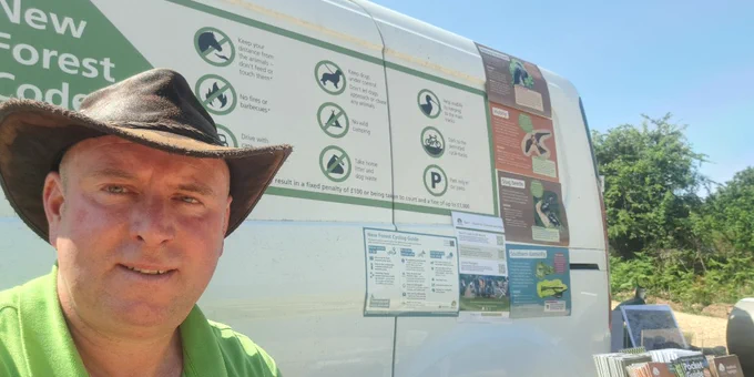 A person wearing a wide-brimmed hat is sitting next to a vehicle displaying informational posters about the "New Forest Code." They are also in front of a table with informational pamphlets. The person is wearing a green polo shirt with "New Forest National Park" branding.