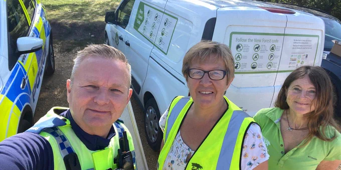A group of three people stand outdoors beside a police car and a van with a "Follow the New Forest code" sign. One person wears a police uniform, and two wear "New Forest" branded vests. Trees and greenery are in the background.