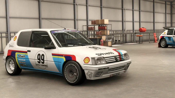 A classic rally car, a Peugeot 205 Turbo 16, is parked indoors. It features a white body with racing decals, including Shell and Michelin logos, and the number 99 prominently displayed. The car is equipped with racing tires and colorful stripes on the side. Another rally car is partially visible in the background within the garage setting.