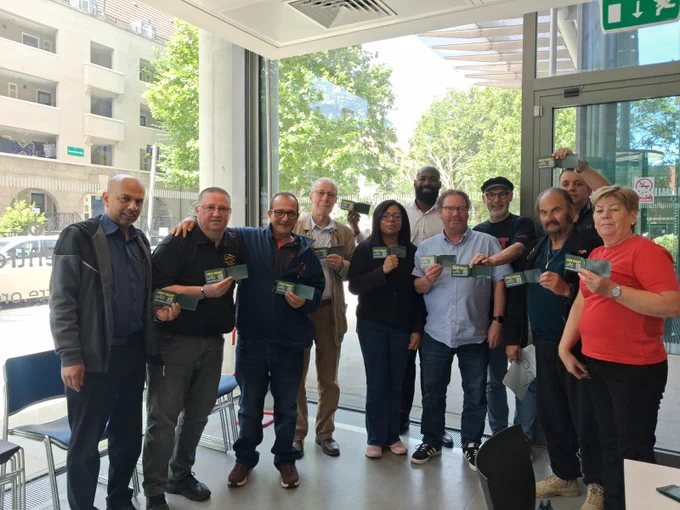 Group photo of RMT members standing in front of large glass windows whilst holding wallets with a card that says ‘Join RMT’.