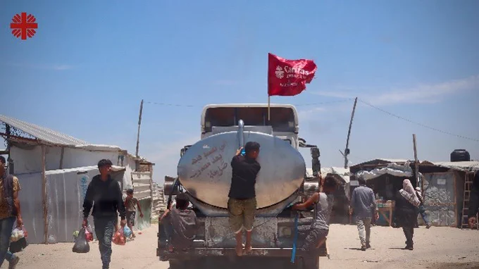 A photo showing a water truck with a Caritas flag driving with a man hanging off the back of it. 