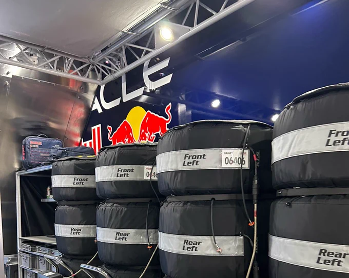 Stacks of Pirelli Formula 1 tyres wrapped in black Oracle Red Bull Racing tyre warmer blankets inside the team garage, labelled “Front Left” and “Rear Left” with driver number 06406. The Red Bull logo and bull mascot are mounted on the wall behind, with overhead lighting rigs above.