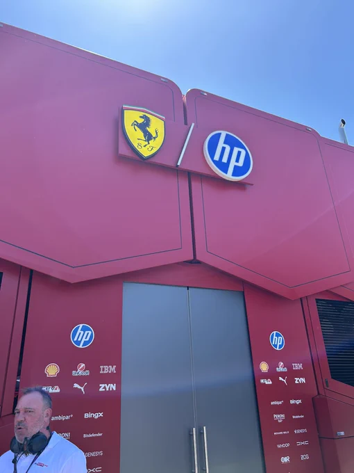 The Scuderia Ferrari HP team building in the Formula 1 paddock under a clear blue sky, with the Ferrari prancing horse and HP logos prominently displayed alongside sponsors including Shell, IBM, Puma, and ZYN. Several people in paddock passes and team kit talk near the entrance.