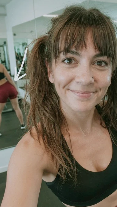 Verschwitzte Frau mit braunem Pferdeschwanz und Pony macht ein lächelndes Selfie im Fitnessstudio